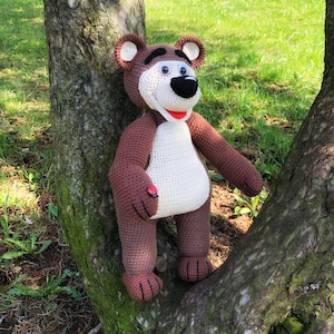 May include: A crocheted brown and white bear with a red nose and black eyes. The bear is sitting on a tree trunk.