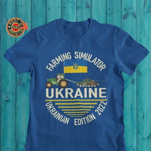 May include: Blue t-shirt with a white circle graphic featuring a tractor pulling a tank, the Ukrainian flag, and the text "Farming Simulator Ukraine Edition 2022".