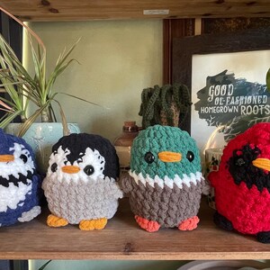 May include: Four crocheted stuffed animals: a blue penguin, a gray penguin, a green duck, and a red cardinal. The stuffed animals are sitting on a wooden shelf. The sign behind the animals reads "Good ol' fashioned homegrown roots."