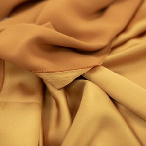 May include: Close-up of a folded piece of fabric in a warm, golden brown color. The fabric has a smooth, silky texture and a subtle sheen.