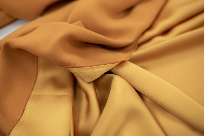 May include: Close-up of a folded piece of fabric in a warm, golden brown color. The fabric has a smooth, silky texture and a subtle sheen.