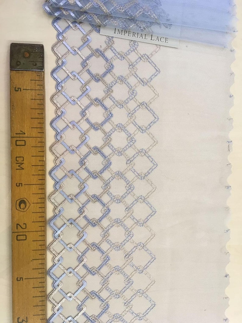 May include: A close-up of blue and silver embroidered lace trim with a geometric pattern. The trim is on a white background and a ruler is visible in the image. The label reads "Imperial Lace".