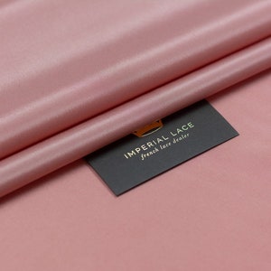 Puede incluir: Primer plano de una tela suave y rosa con un brillo sutil. La tela está doblada por la mitad y una tarjeta negra con letras doradas dice "IMPERIAL LACE french lace dealer".
