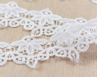 May include: Close-up of white lace trim with a floral design. The lace is intricately detailed with a scalloped edge and is laid out on a light wood surface. The delicate trim is suitable for various craft projects.