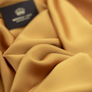 May include: Close-up of a smooth, golden yellow fabric with a subtle sheen. The fabric is draped and folded, creating soft curves and folds. A small black label with the text "WHITE GOLD FABRIC" is visible in the top left corner.