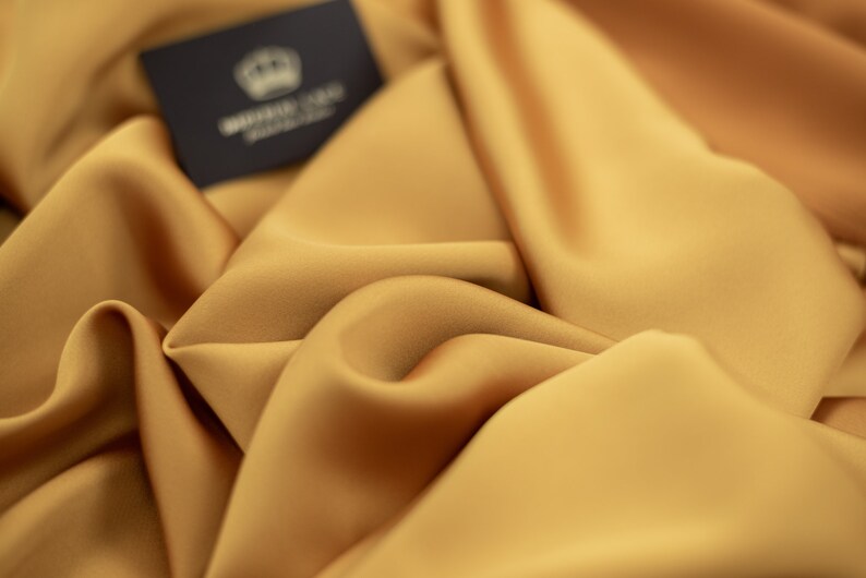 May include: Close-up of a smooth, golden yellow fabric with a subtle sheen. The fabric is draped and folded, creating soft curves and folds. A small black label with the text "WHITE GOLD FABRIC" is visible in the top left corner.