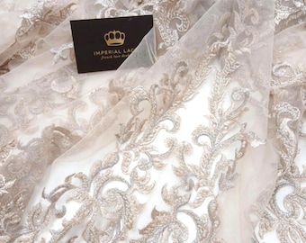 May include: Close-up of beige lace fabric with detailed floral embroidery. The delicate design features silver and gold thread accents, creating a luxurious texture. A business card with a crown logo is visible.