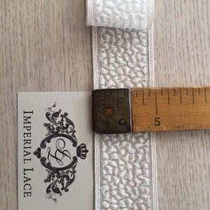 May include: White cotton lace trim with a textured pattern. The trim is approximately 5 inches long and is shown next to a ruler.
