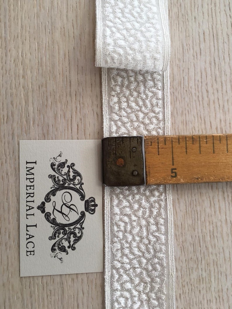May include: White cotton lace trim with a textured pattern. The trim is approximately 5 inches long and is shown next to a ruler.