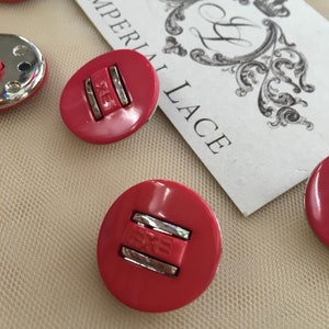 May include: Five pink plastic buttons with silver accents. The buttons have a raised design with the word "EXE" written on them. The buttons are arranged on a white surface with a card that says "Imperial Lace" in black lettering.