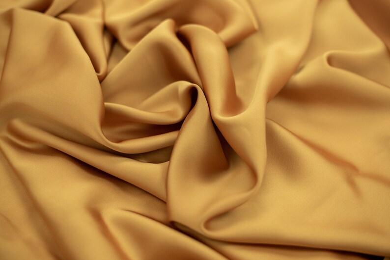 May include: A close-up of a smooth, golden fabric with soft folds and wrinkles. The fabric has a luxurious and elegant appearance.