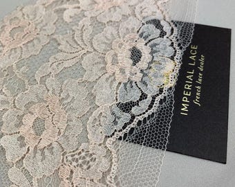 May include: Close-up of a delicate, pale pink lace trim with floral patterns. The lace is intricately designed with a scalloped edge. A black card with the text "IMPERIAL LACE french lace dealer" is partially visible.