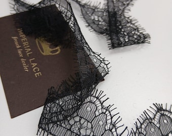 Puede incluir: Cinta de encaje negro con un delicado diseño floral. La cinta se muestra con una tarjeta marrón que dice "IMPERIAL LACE french lace dealer". El encaje tiene un borde festoneado y está sobre una superficie blanca.