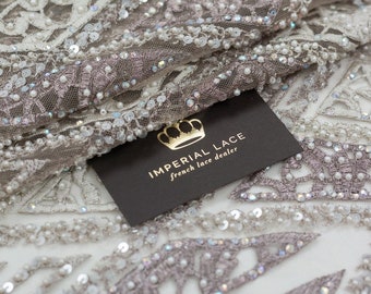 May include: Close-up of a white and silver beaded lace fabric with a geometric pattern. The fabric is draped over a black card with the text "IMPERIAL LACE french lace dealer".