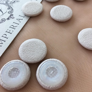 Könnte beinhalten: Nahaufnahme von acht cremefarbenen, stoffbezogenen Knöpfen mit strukturierter Oberfläche. Die Knöpfe sind in einem zufälligen Muster auf einem hellbraunen Netzstoff angeordnet.