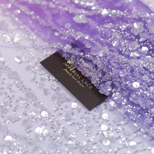Puede incluir: Tela de malla morada con lentejuelas y cuentas plateadas. La tela está etiquetada como "IMPERIAL LACE French lace dealer".