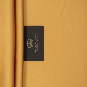 May include: A close-up of a mustard yellow fabric with a black card that says "Imperial Lace French lace dealer" with a gold crown logo.