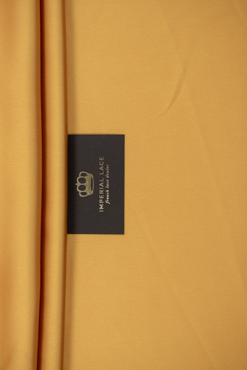 May include: A close-up of a mustard yellow fabric with a black card that says "Imperial Lace French lace dealer" with a gold crown logo.