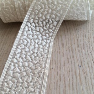 May include: White lace trim with a delicate floral pattern. The trim is rolled up on a wooden surface.