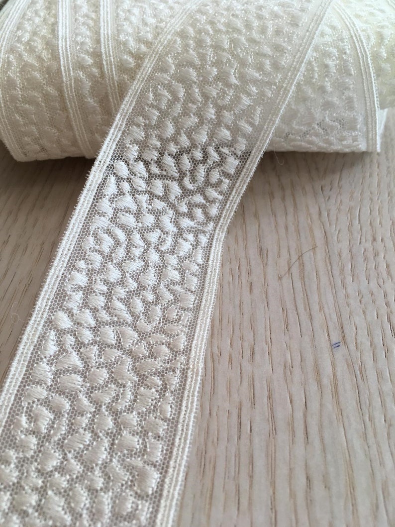 May include: White lace trim with a delicate floral pattern. The trim is rolled up on a wooden surface.