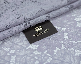 May include: A close-up of a light gray floral lace fabric. The fabric has a delicate pattern of flowers and leaves. The fabric is folded in half, and a black tag with gold lettering reads "IMPERIAL LACE french lace dealer".