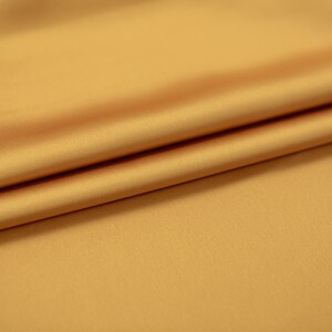 May include: A close-up of a smooth, golden yellow fabric with a soft, silky texture. The fabric is folded in a gentle curve, highlighting its drape and luxurious feel.