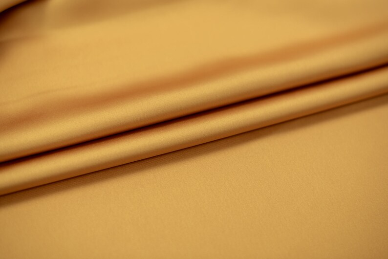 May include: A close-up of a smooth, golden yellow fabric with a soft, silky texture. The fabric is folded in a gentle curve, highlighting its drape and luxurious feel.