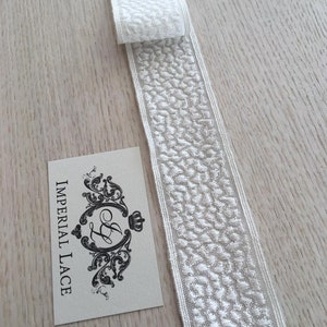 May include: White lace trim with a textured pattern. The trim is folded in half and the label "Imperial Lace" is visible in the background.