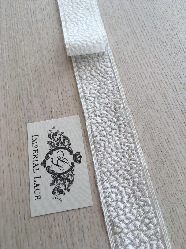 May include: White lace trim with a textured pattern. The trim is folded in half and the label "Imperial Lace" is visible in the background.