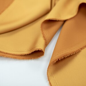 May include: Close-up of a piece of fabric in a warm, golden yellow color. The fabric has a soft, smooth texture and a slightly frayed edge.