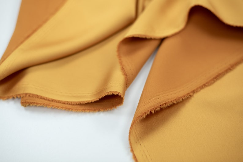 May include: Close-up of a piece of fabric in a warm, golden yellow color. The fabric has a soft, smooth texture and a slightly frayed edge.