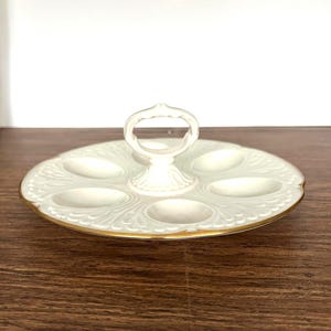 May include: A white ceramic serving tray with a handle, featuring six oval indentations. The tray has a gold trim and a decorative embossed pattern. The handle is arched and the tray is on a wooden surface.