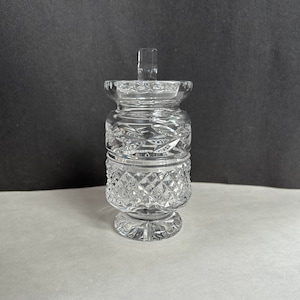 May include: A clear glass jar with a crystal cut design and a removable lid. The jar has a round base and a cylindrical body.