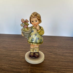 May include: A ceramic figurine of a young girl holding a pot of flowers. The girl wears a yellow dress with blue floral accents and brown shoes. The figurine is on a round, off-white base.