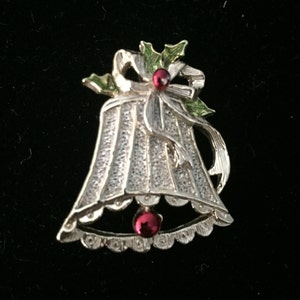 May include: A silver Christmas bell pin with a green holly leaf and red berry accent. The bell has a white textured surface and a scalloped edge.