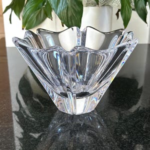 May include: A clear, faceted glass bowl with a starburst design. The bowl has a square base and a scalloped rim. The bowl is sitting on a dark surface, with a plant in the background.