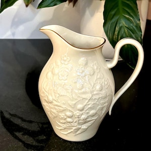 May include: Cream-colored ceramic pitcher with embossed floral design. The pitcher has a curved handle and a gold-colored trim around the rim. The floral pattern includes flowers and berries. The pitcher is likely for serving beverages.