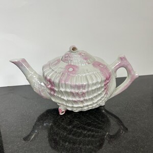 May include: A white teapot with a pink and white shell design. The teapot has a lid and a spout. The teapot is sitting on a black surface.