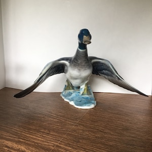 May include: A ceramic duck figurine with outstretched wings, showcasing a detailed design. The duck features a blue head, gray and white body, and dark blue wings. It stands on a blue and white base, with a glossy finish.