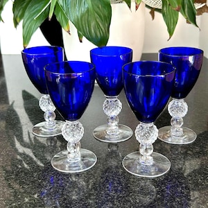 May include: A set of five cobalt blue glass goblets with clear stems. Each goblet features a deep blue bowl and a clear glass stem with a textured, diamond-patterned knop above a circular base. The glasses are arranged in a group.
