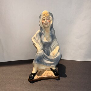 May include: A ceramic figurine of a woman wearing a blue hooded dress and sitting on a base. She has a light complexion and blonde hair.