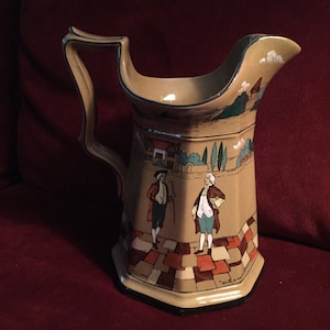May include: A brown and beige ceramic pitcher with a handle. The pitcher has a scene painted on it of two men in 18th-century clothing walking on a cobblestone path. The scene includes a house and trees. The pitcher has a black rim and the words "With a cordial" written on the bottom.
