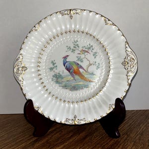 May include: A white decorative plate with a fluted rim and gold trim. The center features a colorful illustration of a bird and a tree. The plate is displayed on a dark brown wooden stand.