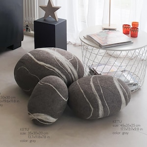 May include: Gray, stone-shaped decorative pillows with white veining. The largest pillow is 23.6x19.6x11.8 inches. A wooden star sits atop a black cube, and a small table holds a lamp and orange candle holders. A cat rests on a black sofa.