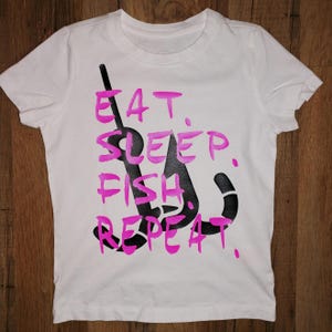 May include: White t-shirt with pink text that reads "Eat. Sleep. Fish. Repeat." and a black fishing hook graphic.