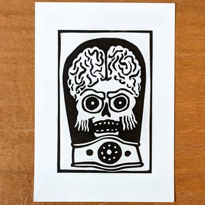 Op de afbeelding: Zwart-wit linoleumsnede van een gestileerde alienkop met een groot brein, grote ogen en een gesegmenteerde nek. De prent staat op wit rechthoekig papier met een zwarte rand.