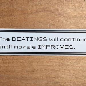 May include: A white sticker with a black border and the text "The BEATINGS will continue until morale IMPROVES." The sticker is rectangular and has a pixelated design, set against a wooden background.