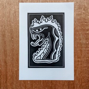 May include: Black and white linocut print of a snake with flames around its head. The snake is facing the left side of the image and has a large, open mouth with sharp teeth. The flames are stylised and have a jagged, pointed shape.