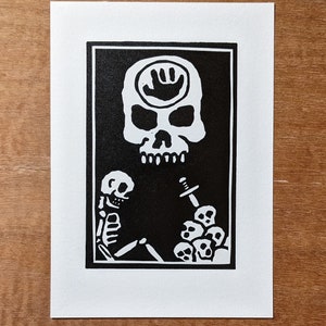 Op de afbeelding: Zwart-witte linosnede van een schedel met een handafdruk symbool op zijn voorhoofd. De schedel is omringd door een rand van kleinere schedels en een zwaard.