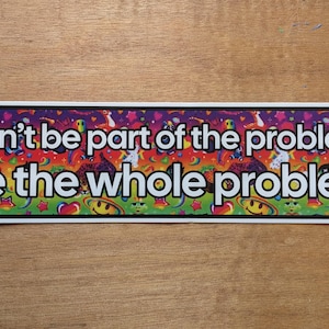 May include: A rectangular bumper sticker with a white border and a vibrant, multicoloured background featuring various small icons like smiley faces, stars, and planets. The text reads in bold white letters: Don't be part of the problem, be the whole problem.